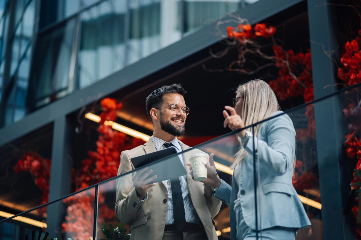 Two business colleagues are standing on a balcony, having a conversation about work while holding a tablet and a coffee cup